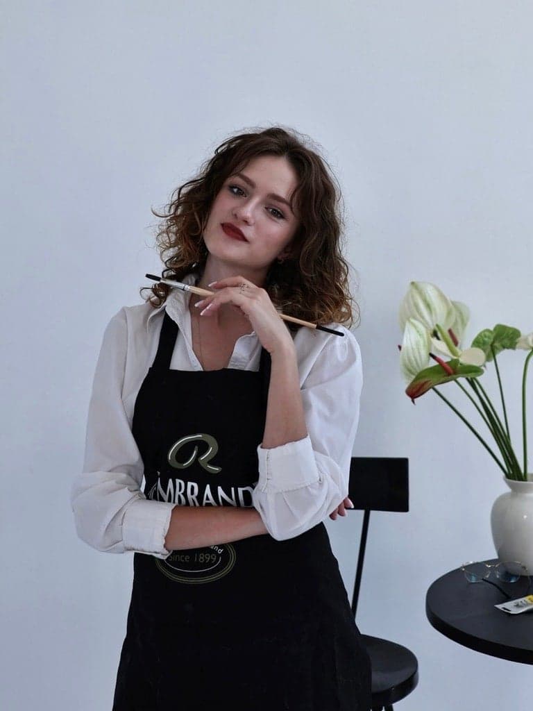 Angelina Ogul w czarnym fartuchu malarskim, z pędzlem uniesionym przy twarzy, w pracowni