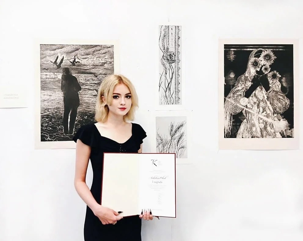 Angelina Ogul na wystawie, z dyplomem, na tle swoich prac graficznych